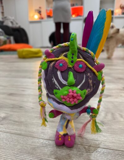 Personnage modelé violet, orné de fils et de plumes colorées.